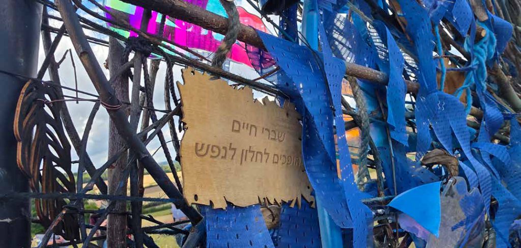 Holzplakette mit hebräischer Inschrift zwischen blauen Stoffstreifen